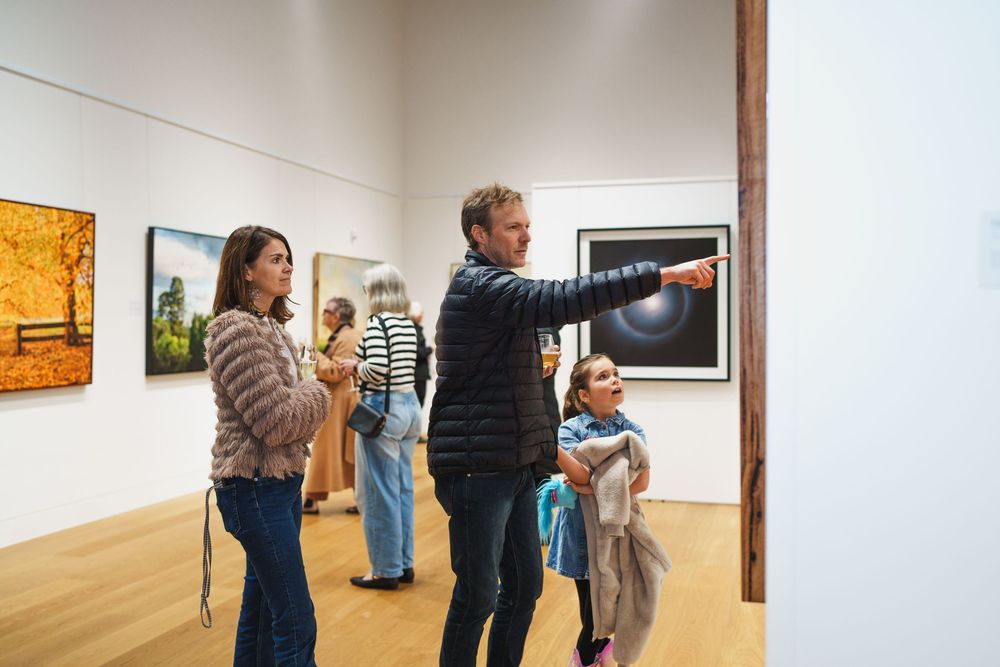 Family exploring art gallery, man pointing at large circular eclipse photograph on white wall.