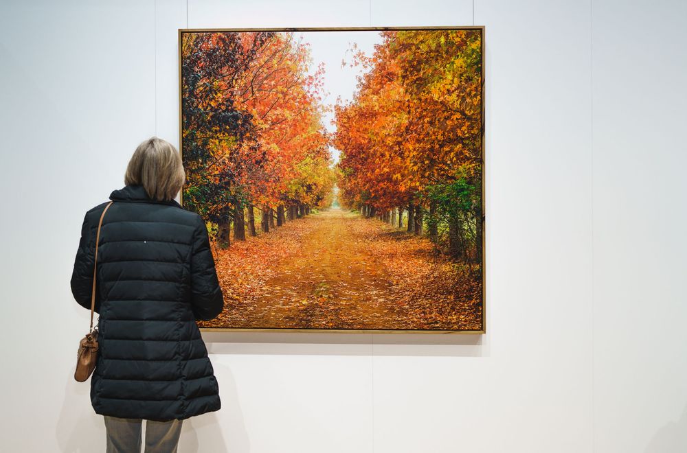 Woman admiring vibrant autumn tree-lined pathway painting in a well-lit art gallery.