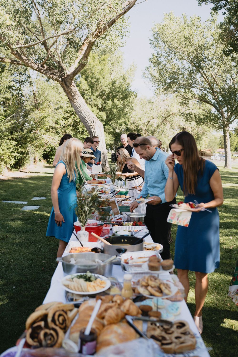 Guests serve themselves at an outdoor wedding buffet table laden with food, set on green grass under trees.
