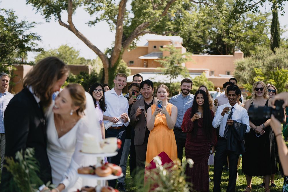 Wedding guests applaud and smile while watching the couple cut their cake at an outdoor garden reception.