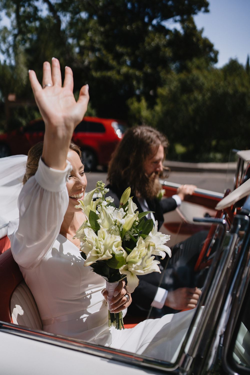 Smiling bride waves while holding white lily bouquet seated in a classic convertible car on her wedding day.