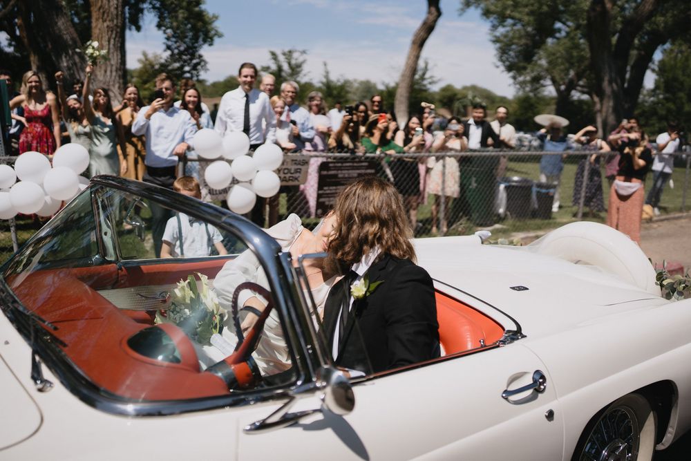 Newlyweds depart in a vintage white convertible as guests cheer and wave with white balloons at outdoor wedding at a park in Albuquerque