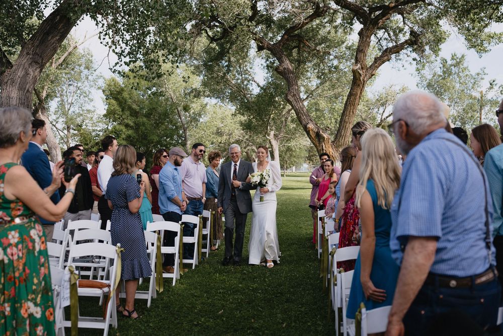 Bride walking down outdoor wedding aisle with guests seated in white chairs under large shade trees in a public park in Albuquerque