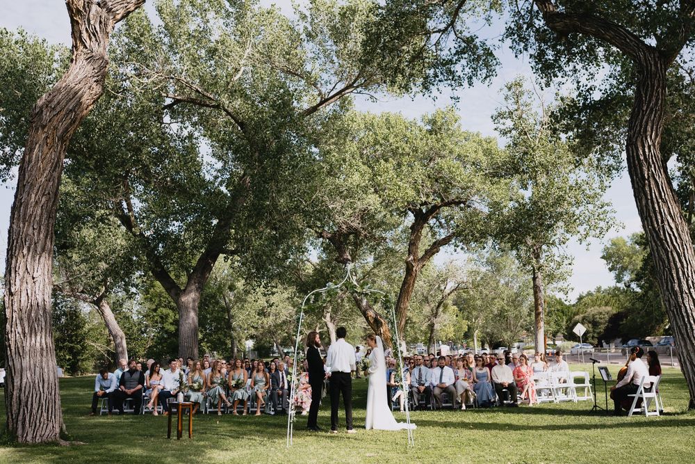 Outdoor wedding ceremony under sprawling oak trees with guests seated in white chairs on green lawn.