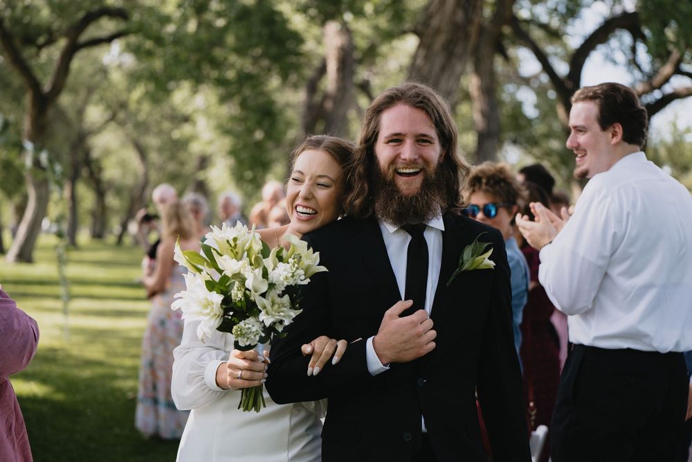 Joyful newlywed couple walking back down the aisle after outdoor wedding ceremony holding white flowers.