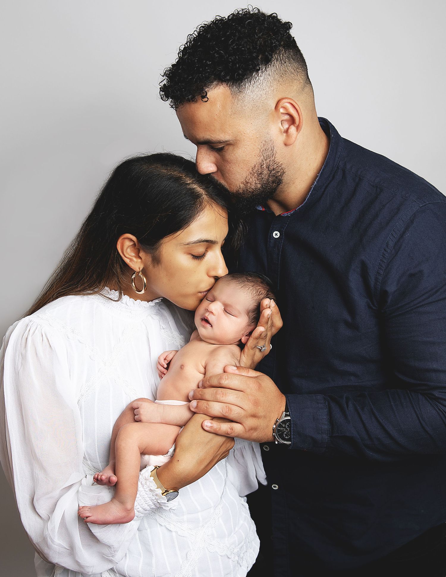 Young couple tenderly kissing their newborn baby, mother in white dress, father in navy shirt. During a newborn photoshoot in Sidcup
