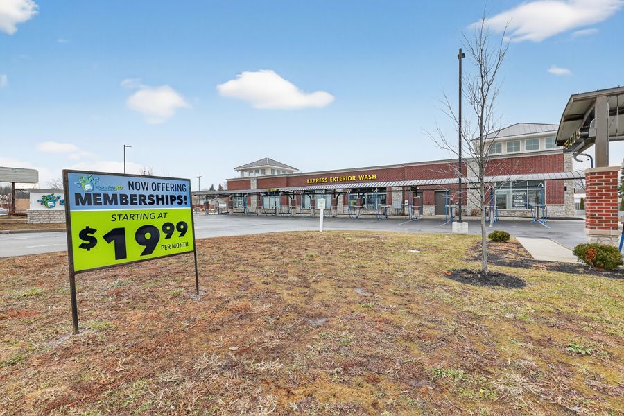 Strip mall with membership sign starting at $19.99, featuring brick storefronts, young trees, and green lawn under cloudy sky.