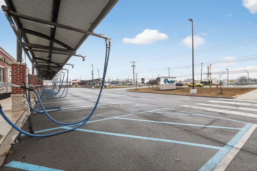 Outdoor car wash station with blue vacuum hoses hanging under a metal canopy beside an empty parking lot.