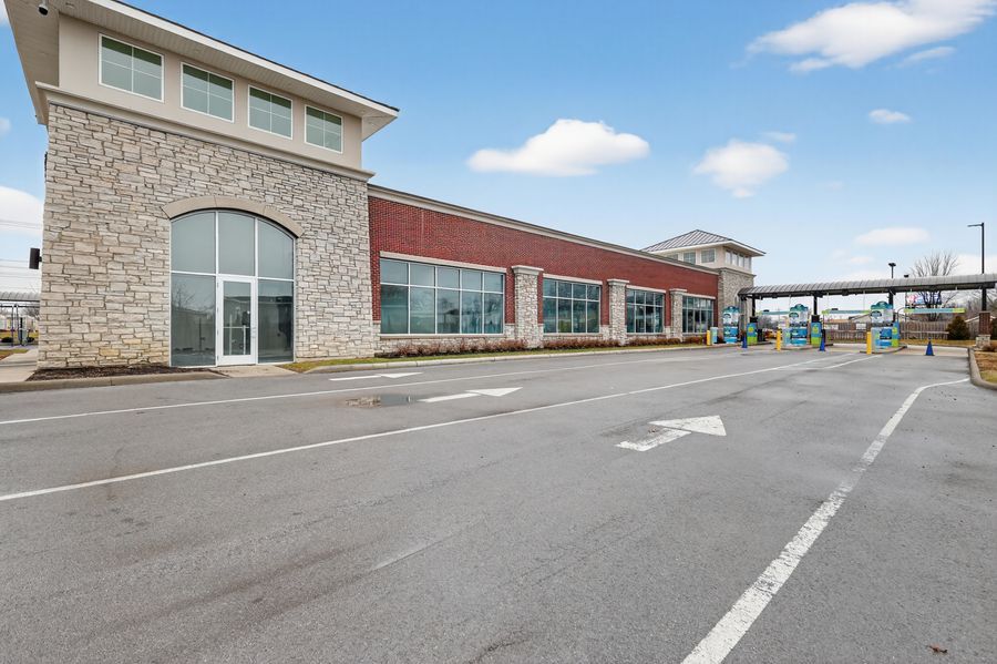 Modern retail strip mall with stone and brick facade, large windows, and spacious parking lot under a partly cloudy sky.