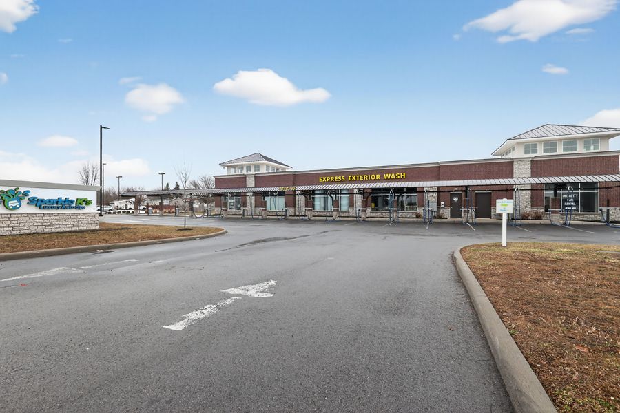 Spacious strip mall parking lot with retail stores including a green-signed shop under a partly cloudy sky.