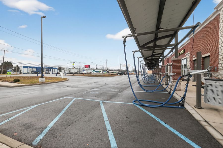 Empty car wash facility with blue vacuum hoses under a covered canopy and marked parking spaces in a commercial lot.