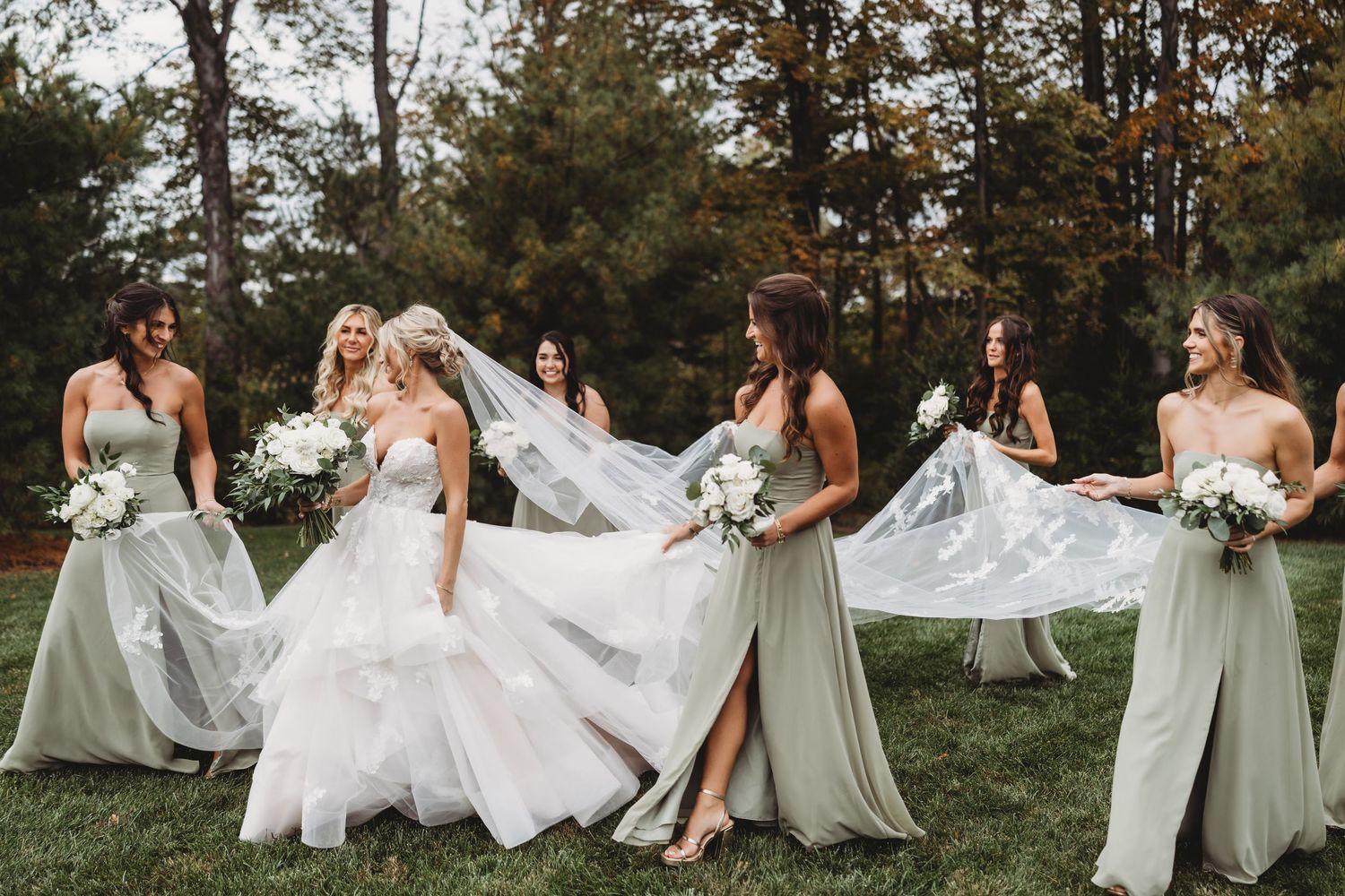 Bride in white gown with bridesmaids in sage green dresses holding bouquets on a lush green lawn.