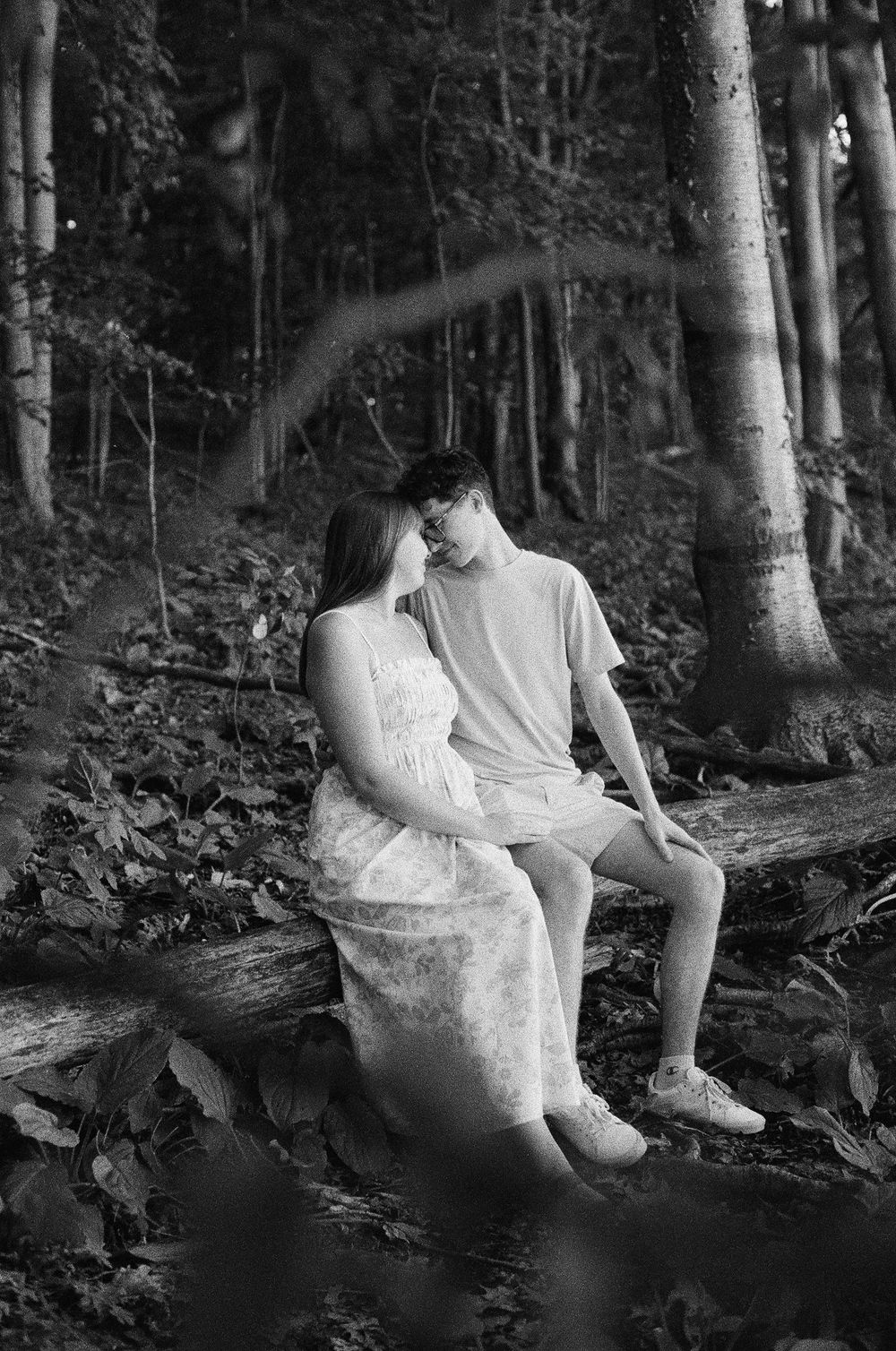 Black and white photo of a young couple sitting on a fallen log in a forest, leaning close together romantically.