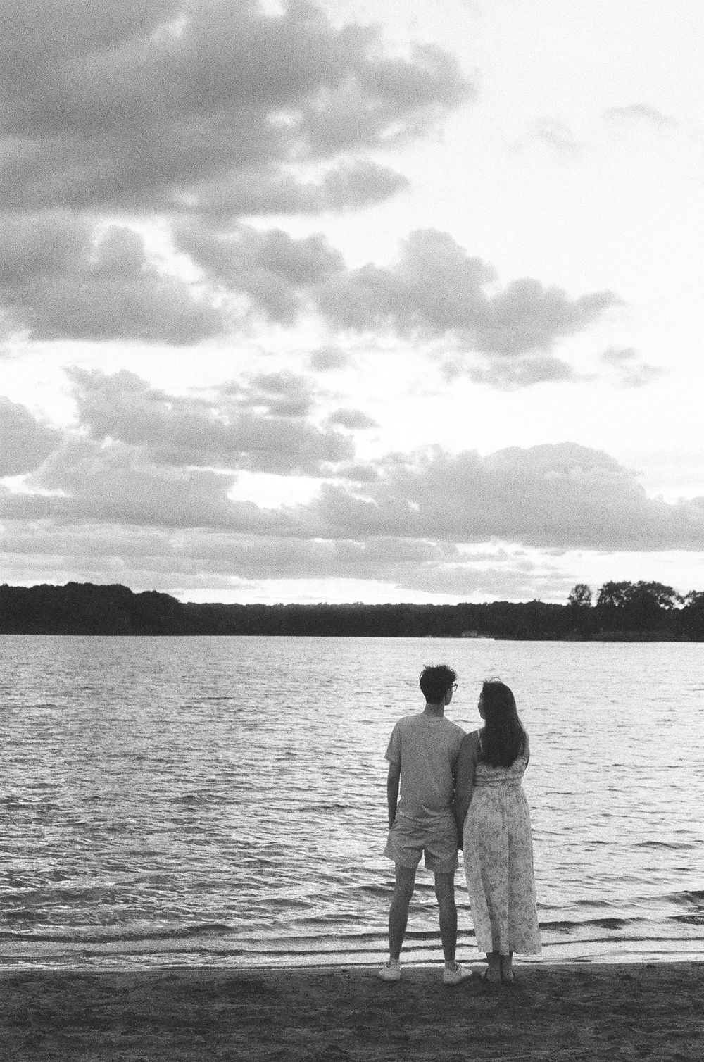 Black and white silhouette of a couple standing together on a beach gazing at a cloudy sky.