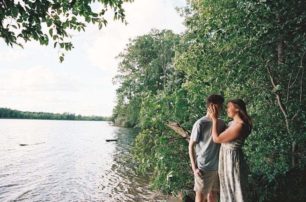 Couple standing by a serene lakeside surrounded by lush green trees, pointing at nature.