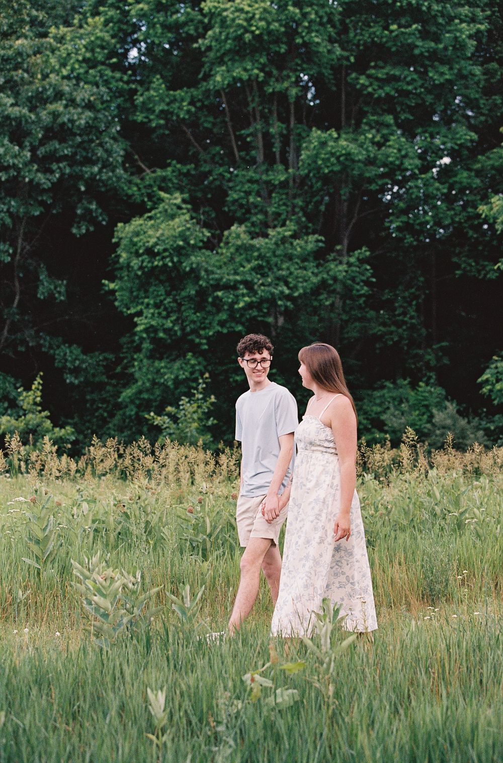 A couple holds hands in a lush green meadow surrounded by tall trees on a warm summer day.