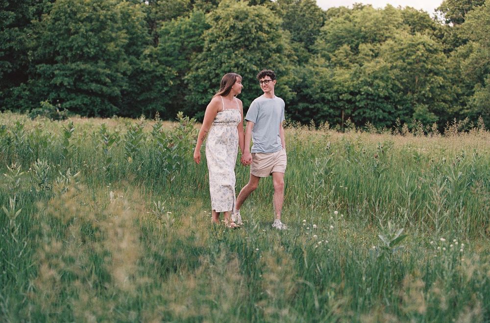 A couple walks hand in hand through a lush green meadow with tall grass and trees in the background.