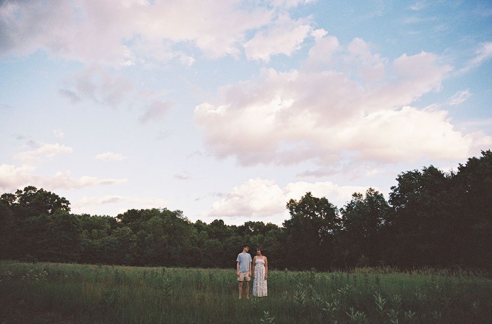 Couple standing together in a lush green meadow at dusk with dramatic cloudy sky and tree line in background.
