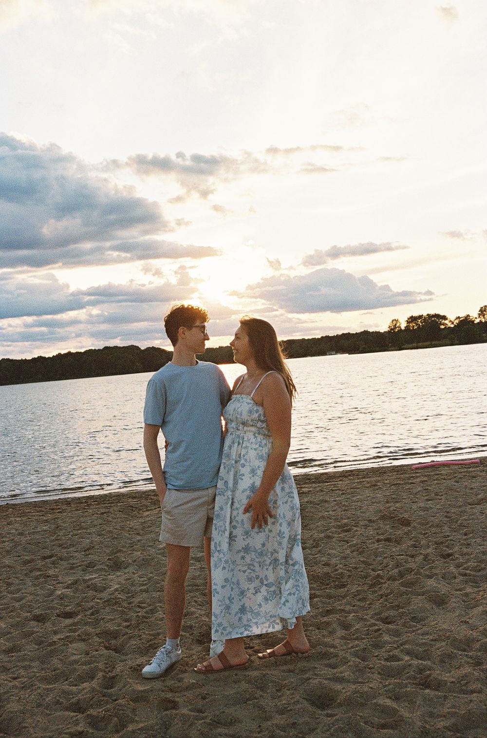 A young couple stands together on a sandy beach at sunset, smiling at each other near a calm lake.