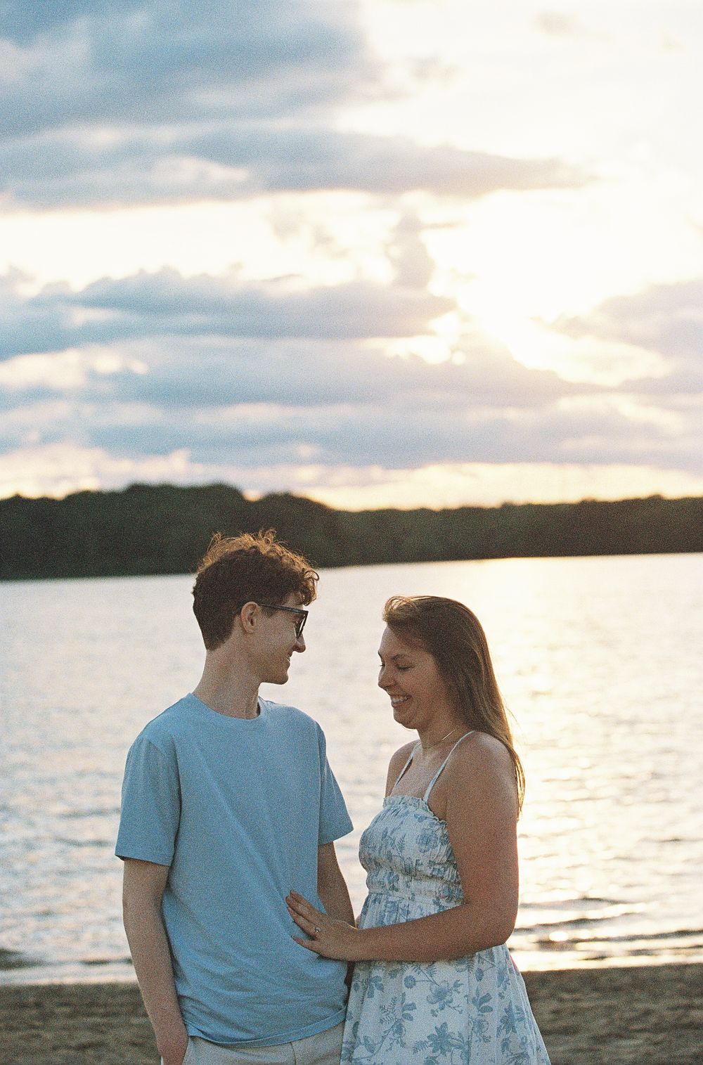 Smiling couple holding each other close on a beach at sunset beside a calm lake.
