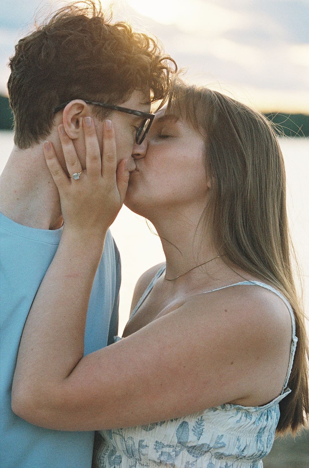 A young couple shares a romantic kiss outdoors near a lake at sunset, woman wearing a ring and floral dress.
