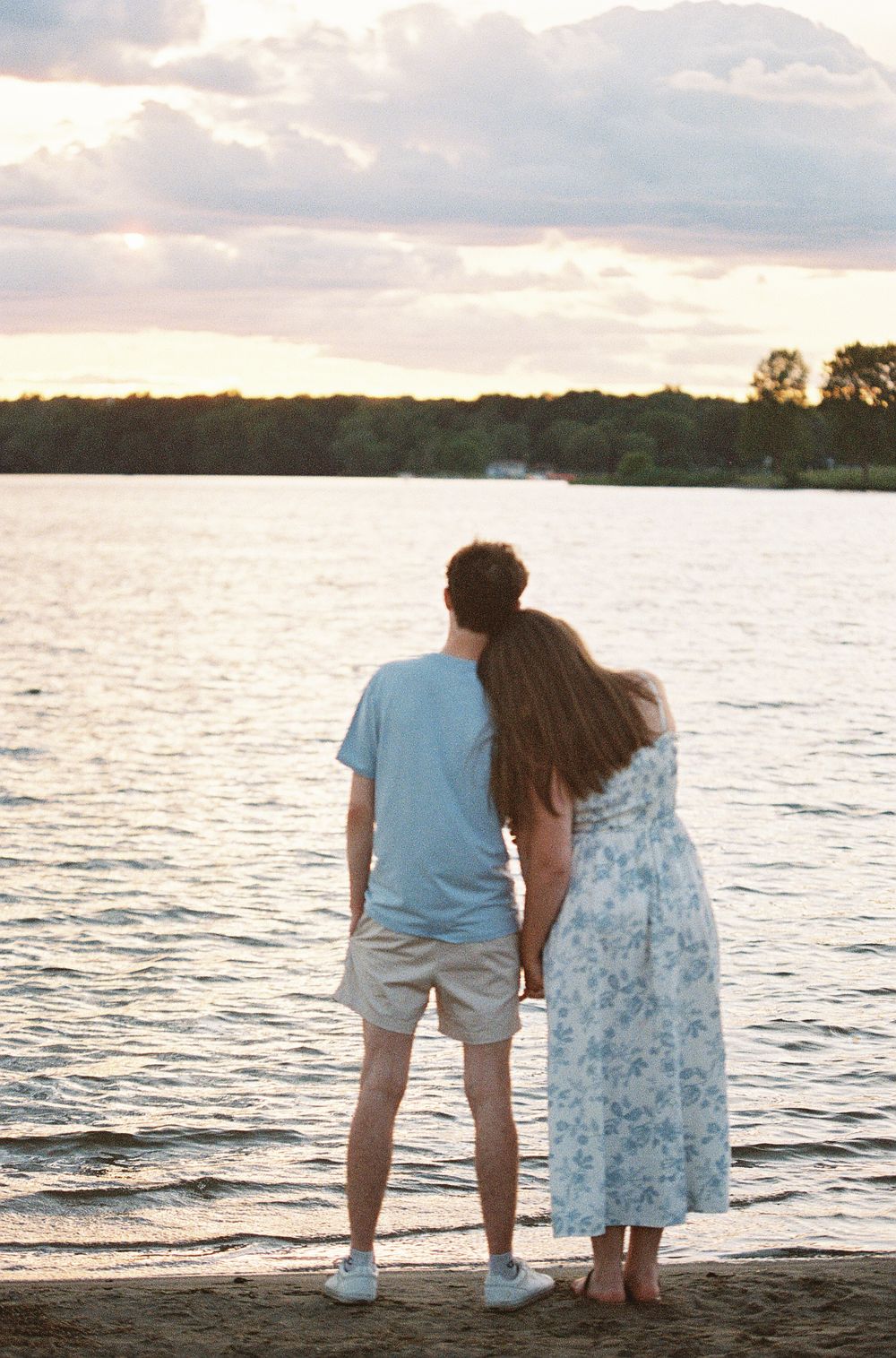 A romantic couple holds hands while gazing at a serene lake during sunset, woman resting her head on mans shoulder.