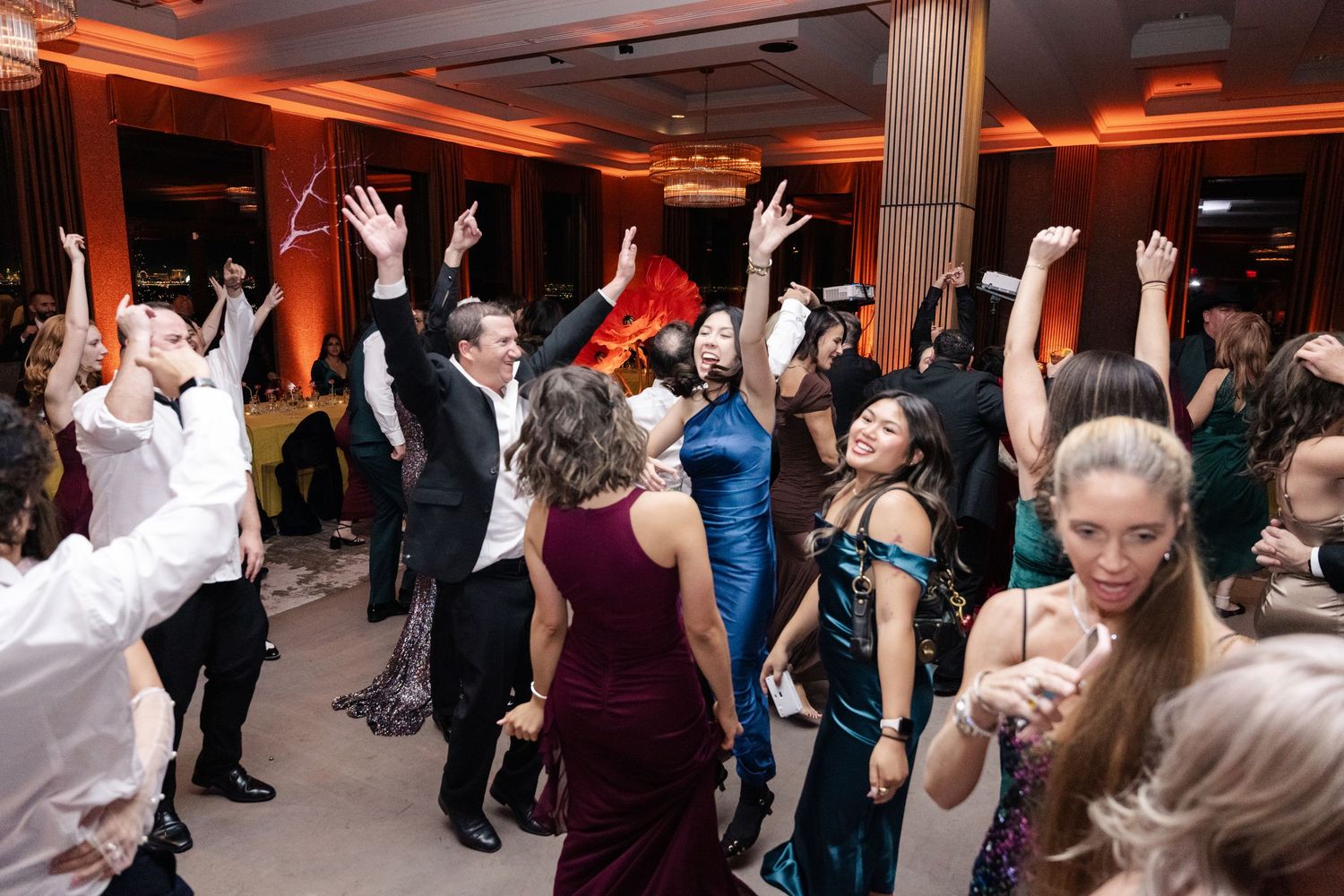 Guests dancing energetically at a formal event, wearing elegant attire in a warmly lit ballroom.