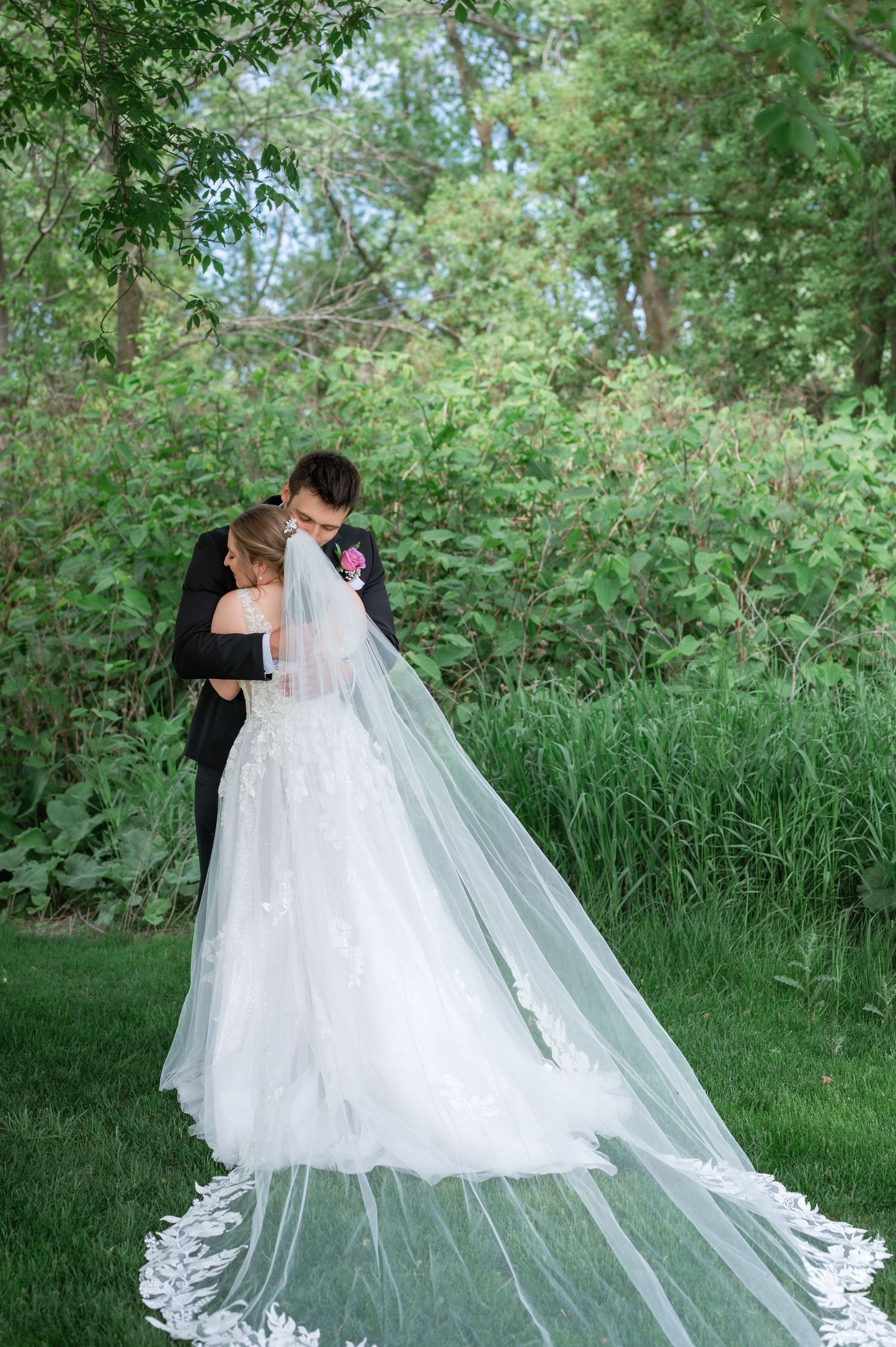 A bride in a flowing white gown with cathedral veil embraces her groom in a lush green garden setting.