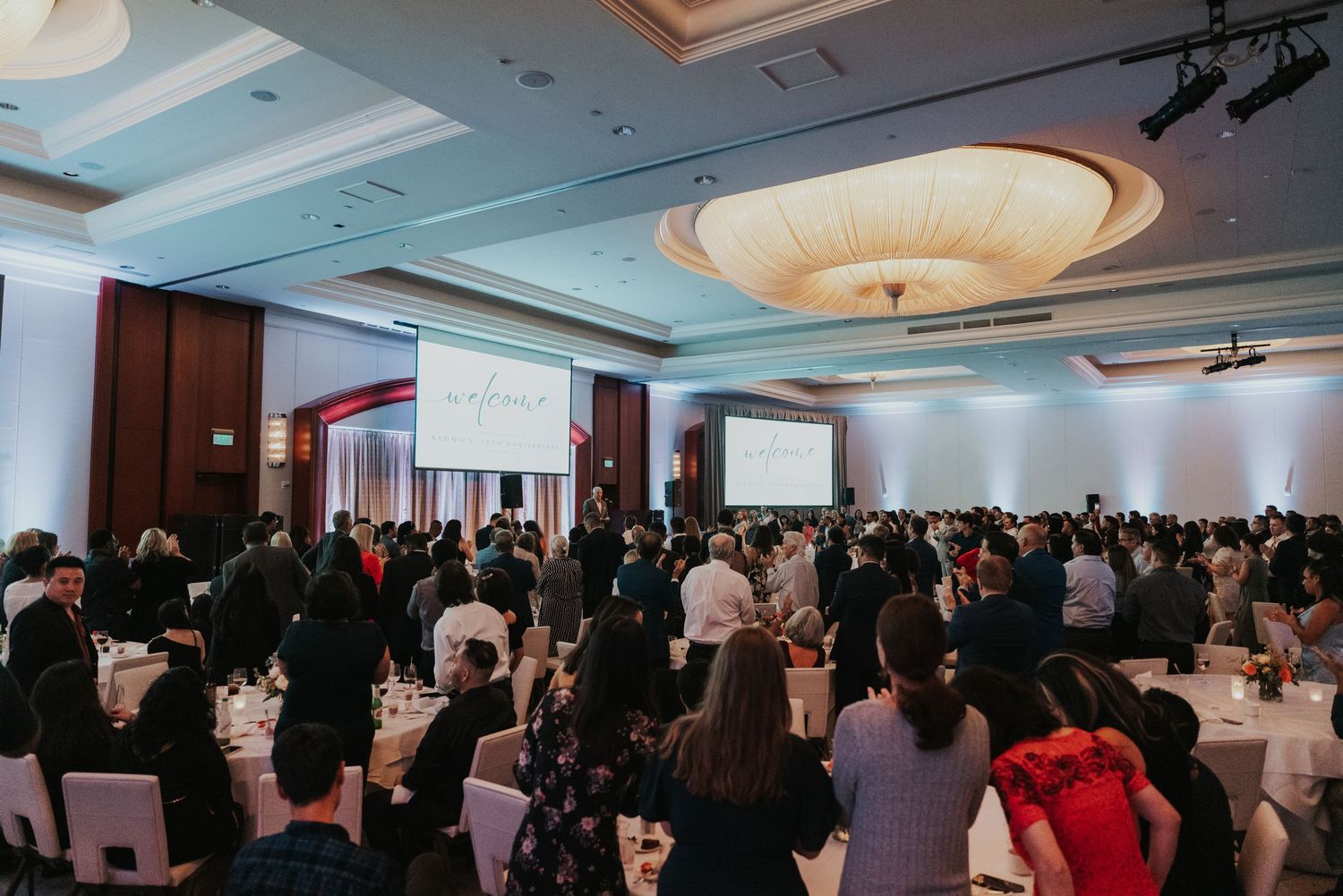 Large corporate gala event in elegant ballroom with guests standing at round tables beneath ornate ceiling lights.