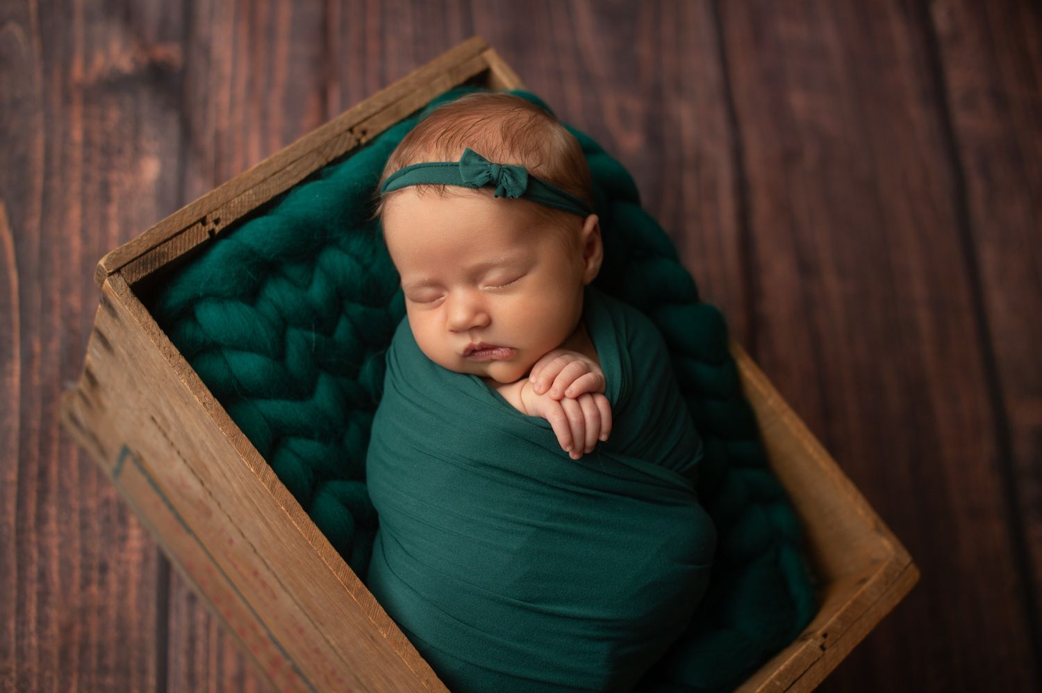 Sleeping newborn baby swaddled in teal green wrap lying in rustic wooden crate with matching knit blanket.