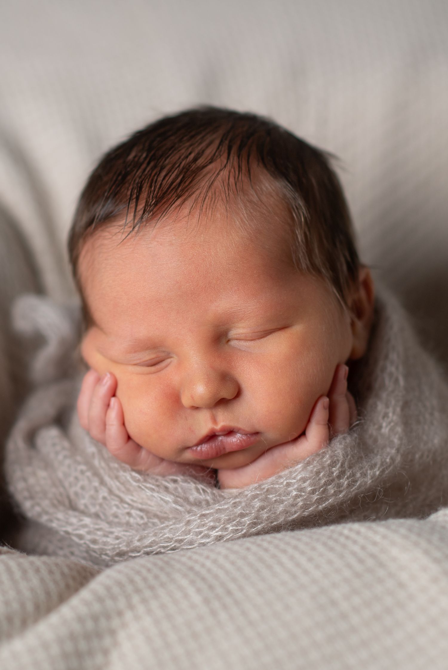 Sleeping newborn baby wrapped in soft gray knit swaddle with hands tucked under cheeks in a peaceful pose.