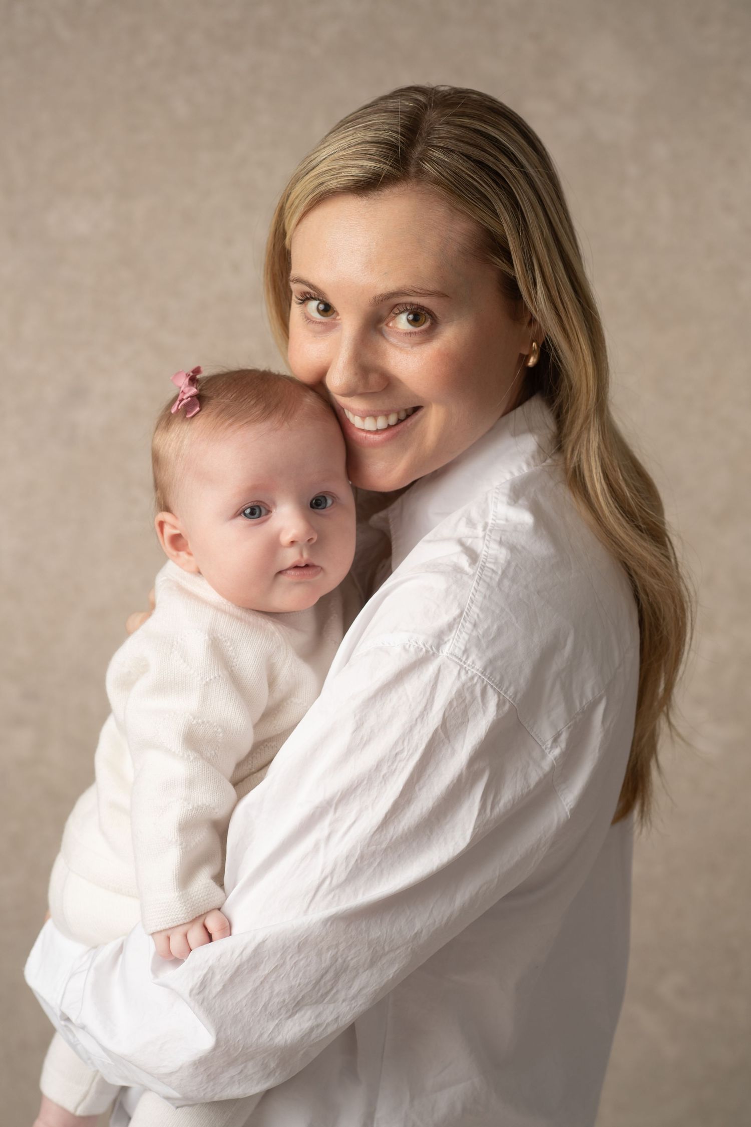 Smiling blonde woman in white shirt holding a baby girl with a pink bow in a studio portrait.
