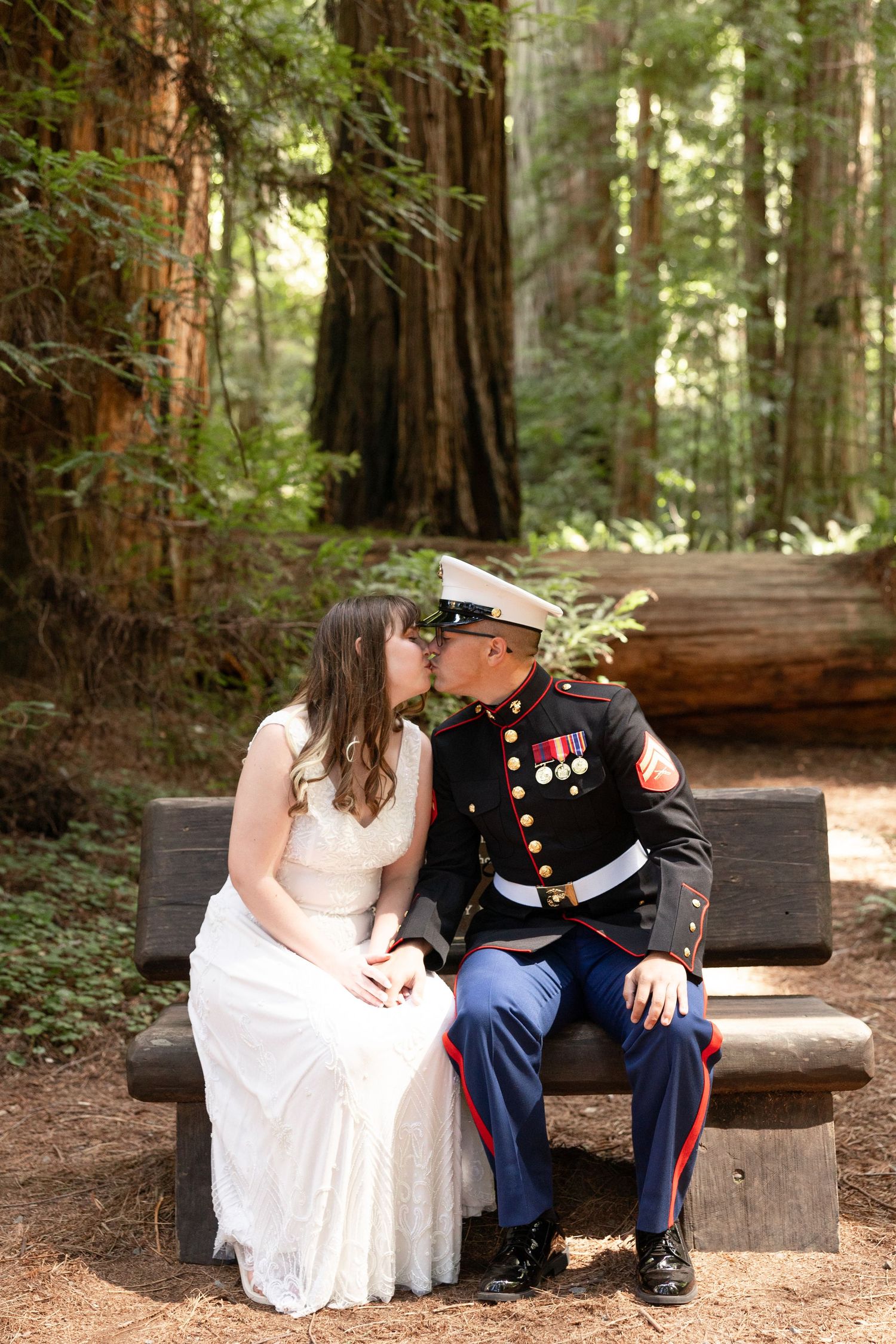 Marine groom and bride share a kiss among towering redwood trees in a sunlit forest wedding ceremony.
