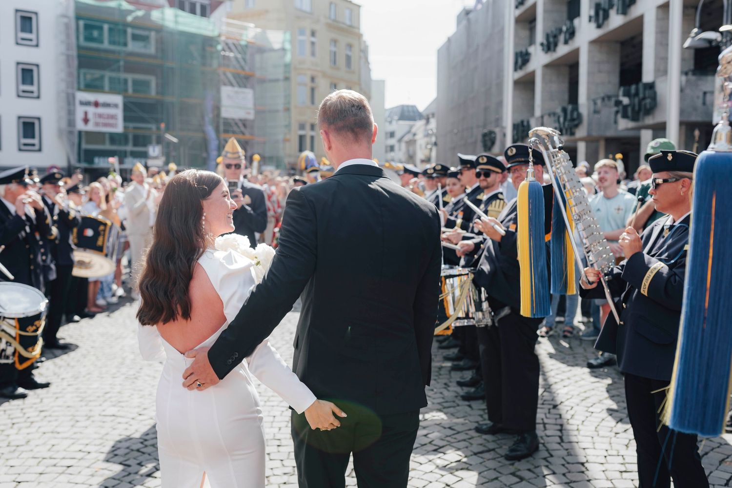 Bride and groom walk hand in hand past a marching band during their outdoor wedding ceremony.