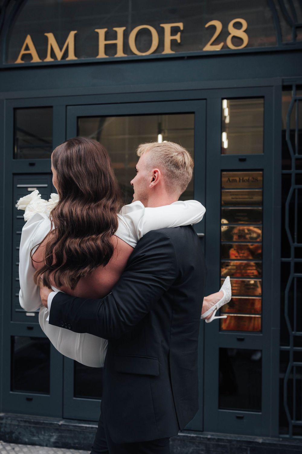 A couple embraces in wedding attire outside Am Hof 28 building, bride in white gown and groom in dark suit, rear view.