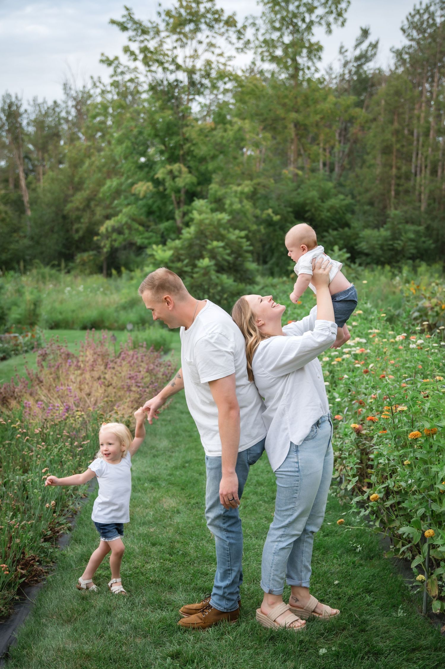 A young family poses in a lush garden, parents in white tops and jeans holding two small children outdoors.