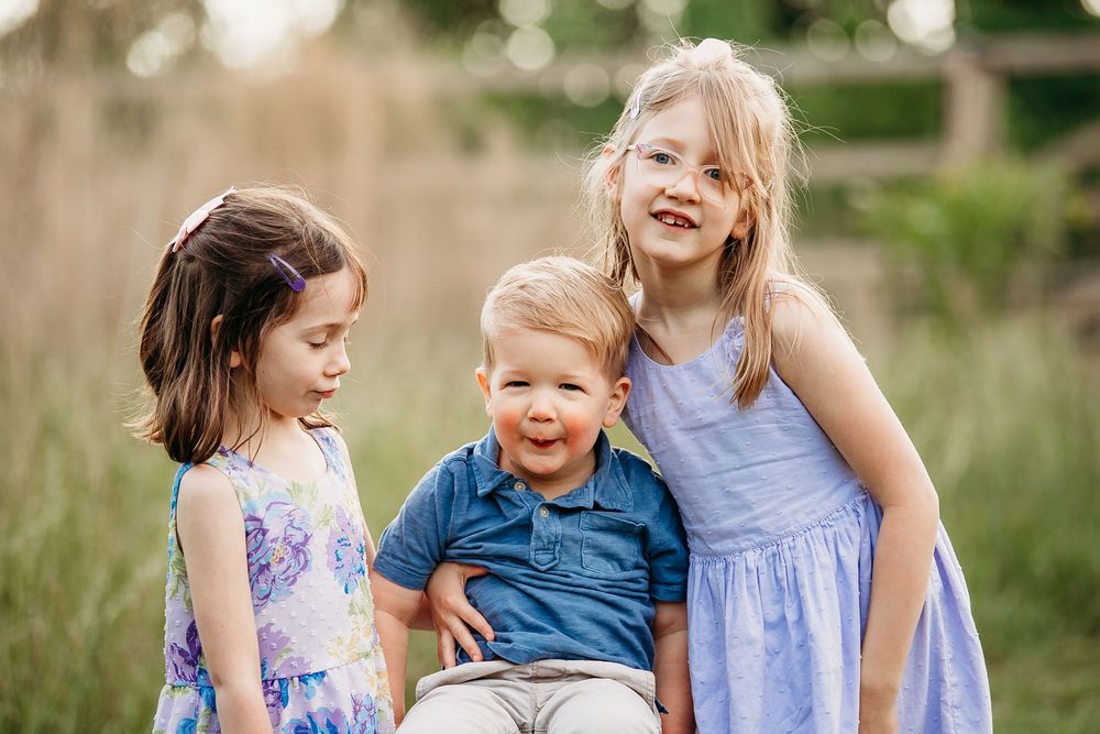 Three young siblings smiling and hugging together in sunny outdoor field setting.