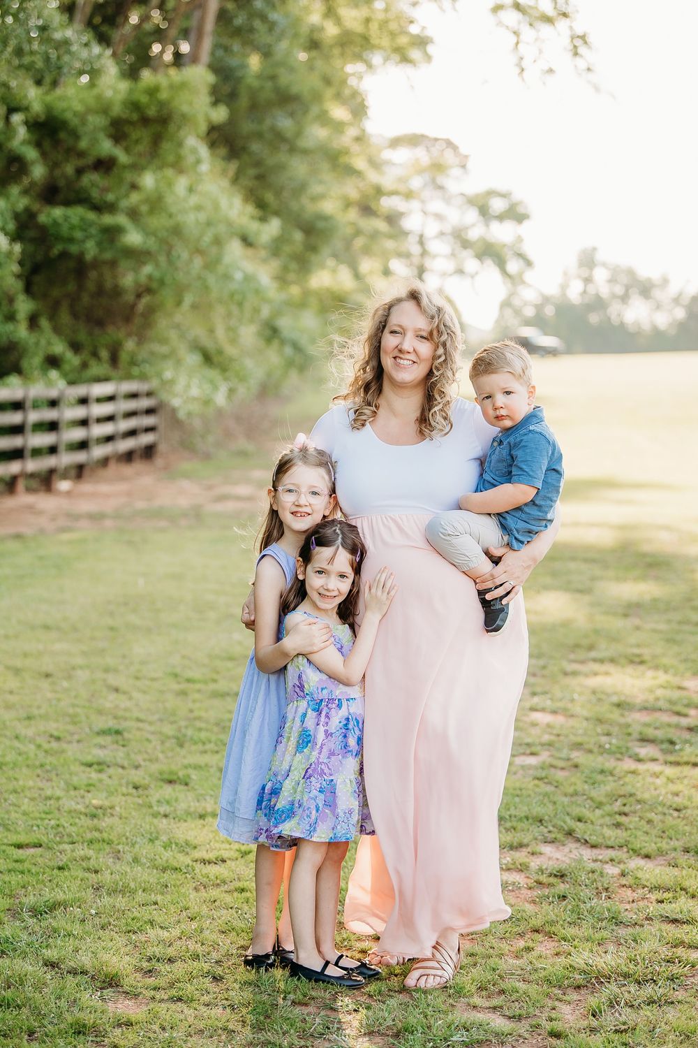 Curly-haired pregnant mother holding toddler son while two daughters hug her outdoors.