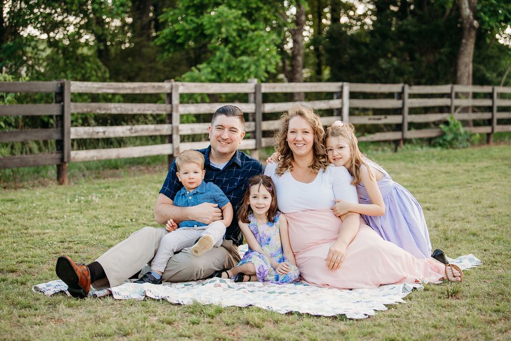 Happy family of six sitting on blanket in grassy field near wooden fence outdoors.