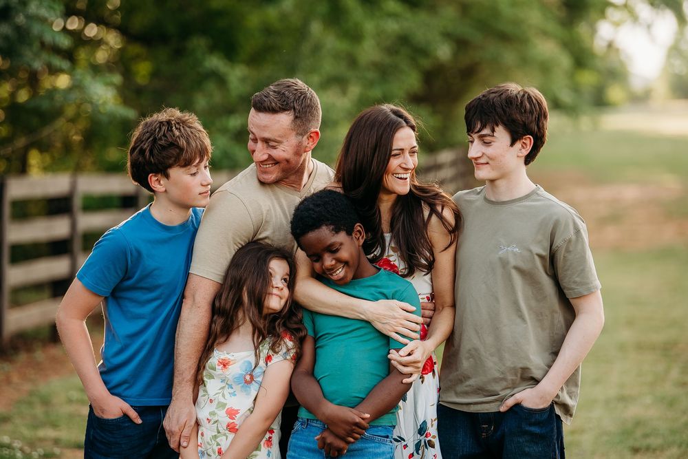 Happy family of six smiling and embracing outdoors in a green field during a warm summer day.