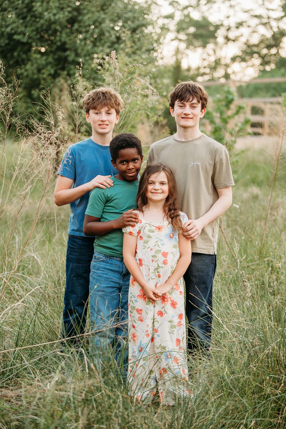 Four siblings posing together in a tall grass field, smiling for a family portrait outdoors.