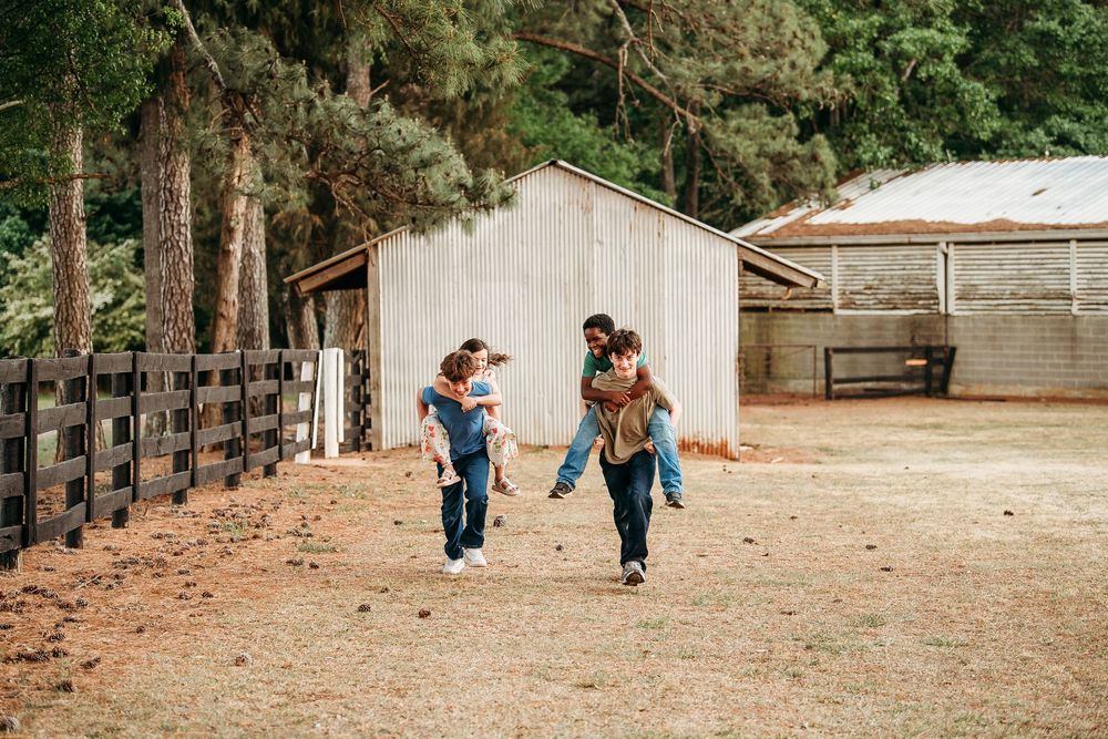 Family walking together near a barn, with parents giving children piggyback rides outdoors.