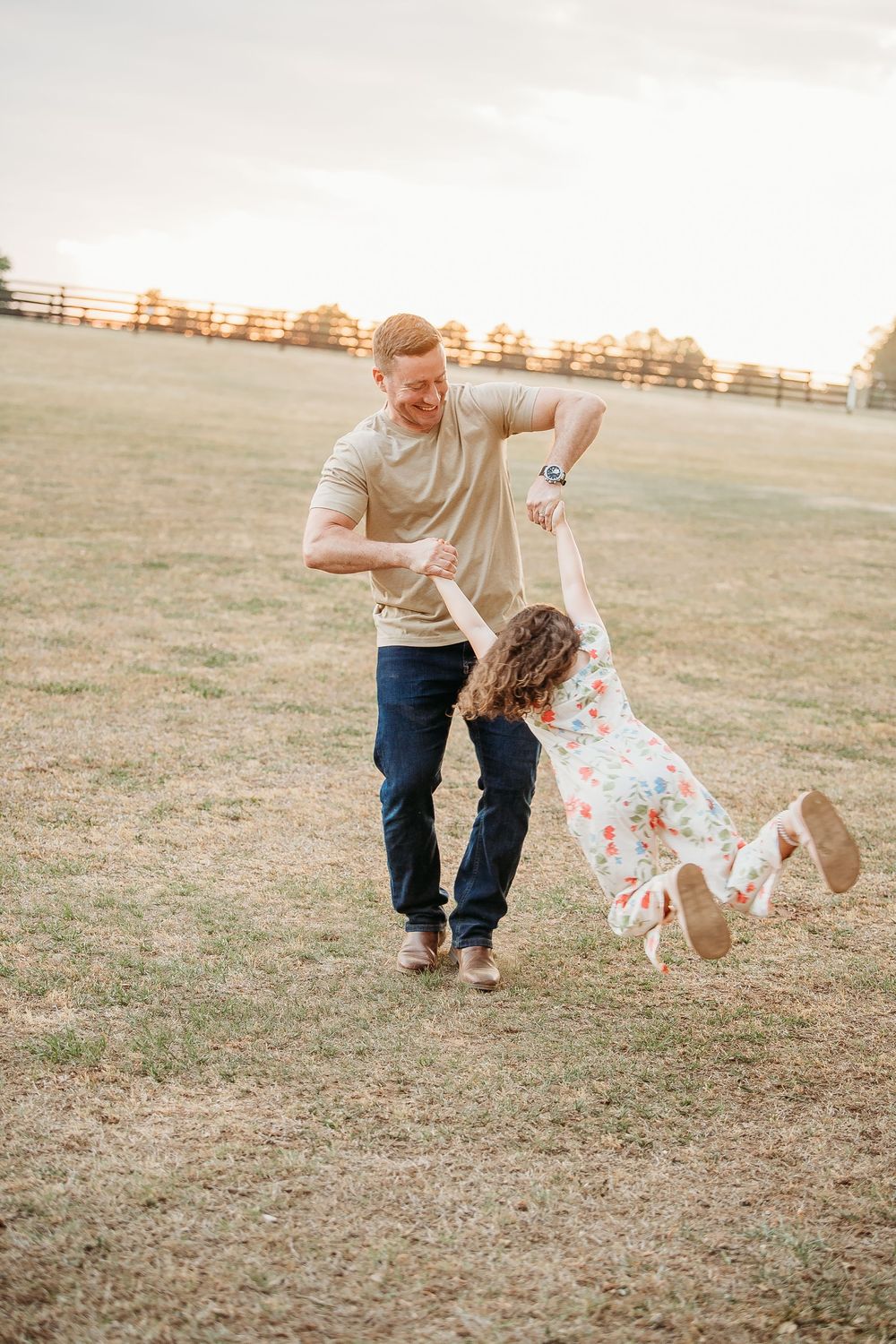Father joyfully spinning his young daughter in a floral dress in an open grassy field at sunset.