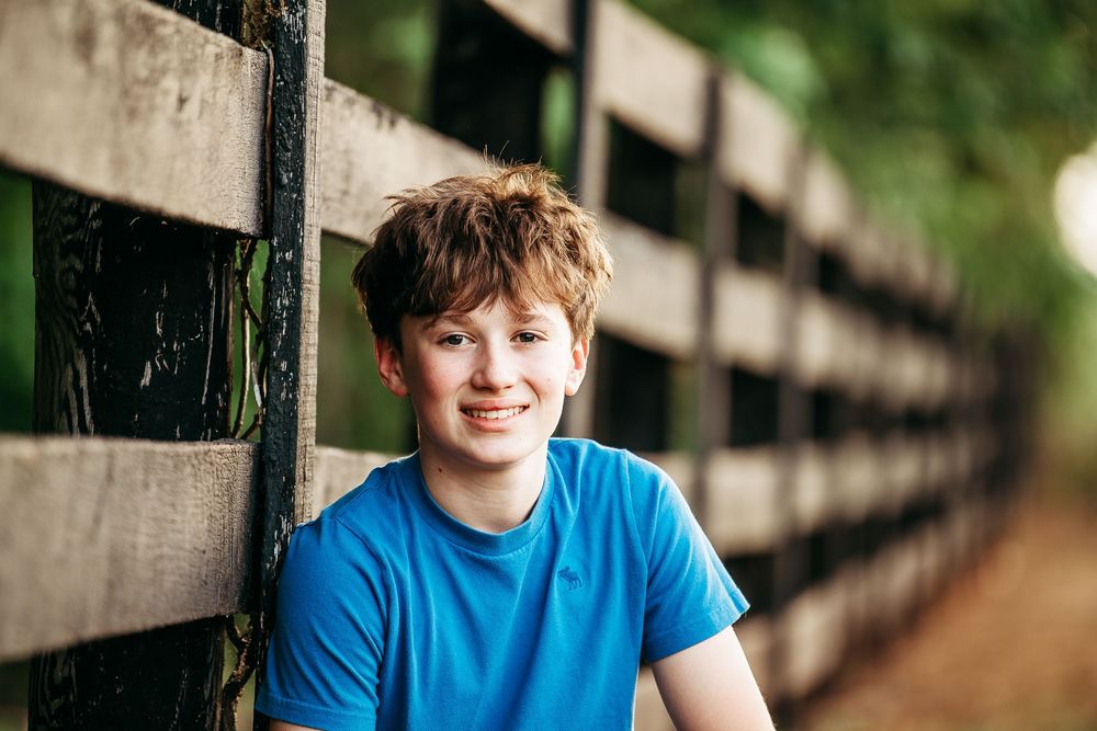 Smiling teenage boy with red hair wearing a blue shirt leaning against a wooden fence outdoors.
