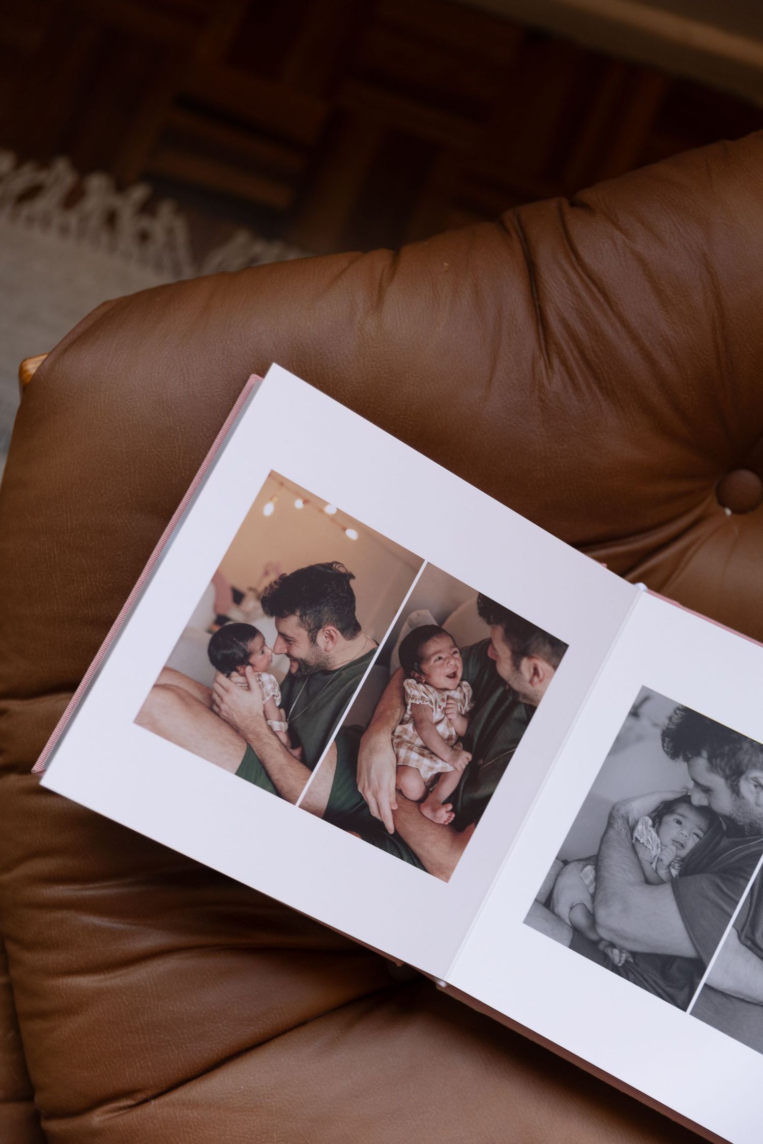 Open photo album on brown leather couch displaying newborn baby with father in warm family portrait photos.