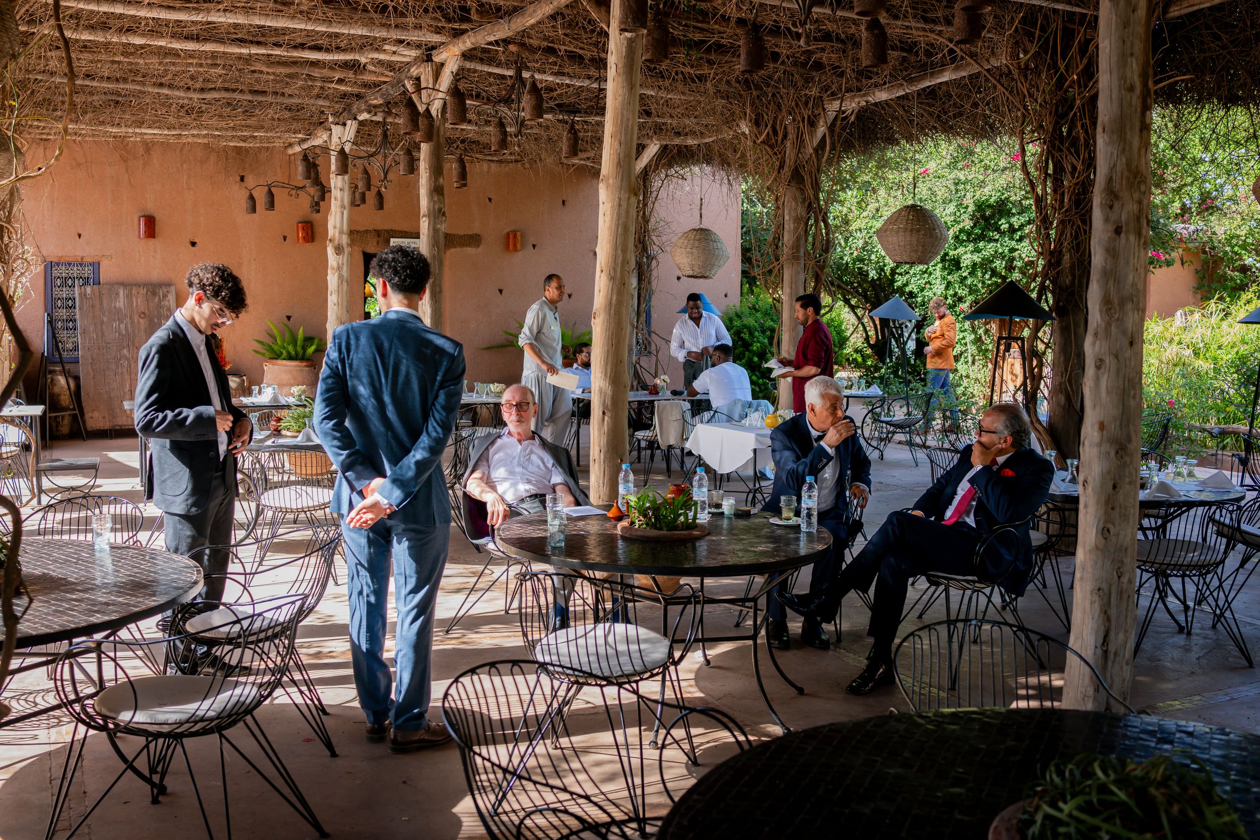People socializing on a rustic outdoor terrace with iron chairs and tables, surrounded by lush greenery and warm earthy walls.