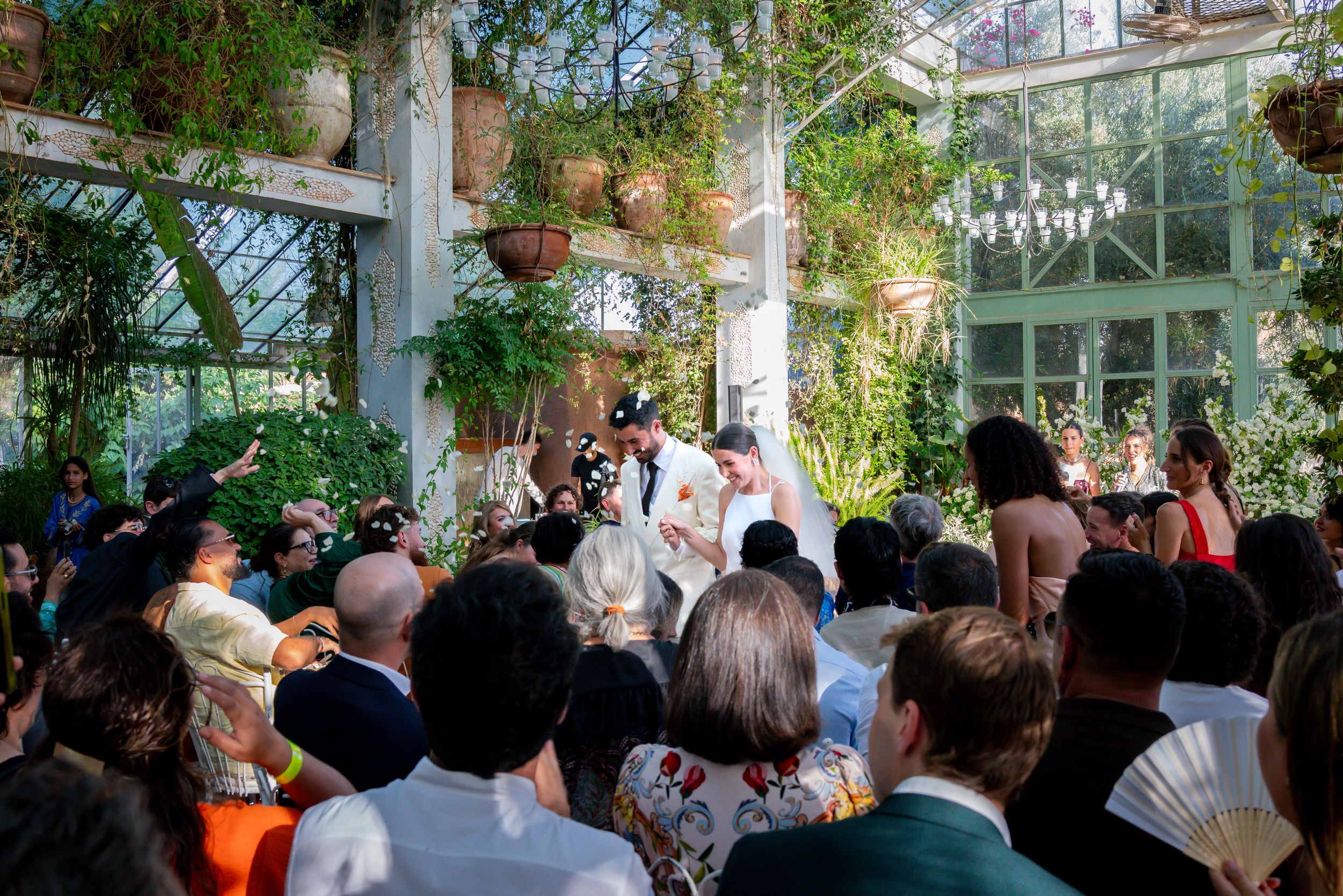 Romantic wedding ceremony in a lush greenhouse with guests surrounding the couple amid tropical plants and natural light.