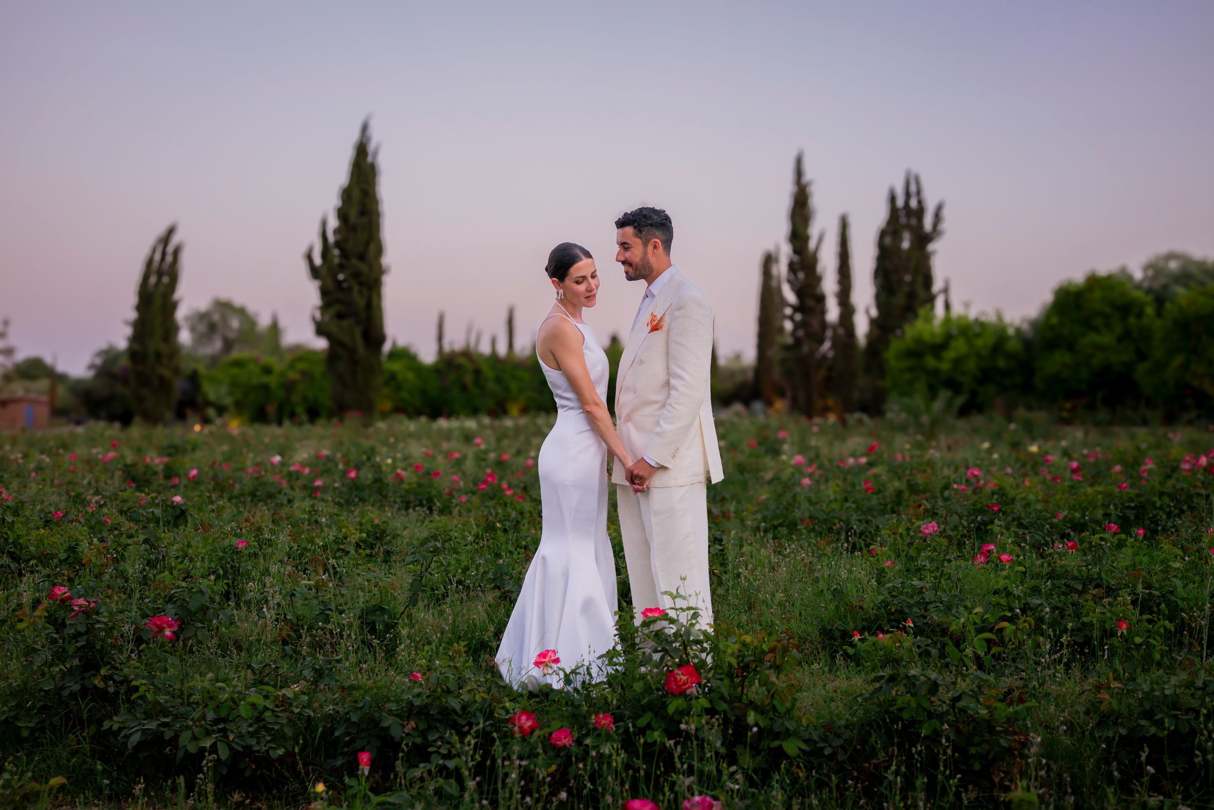 Bride and groom standing in a lush flower field at dusk, sharing a romantic moment in wedding attire.