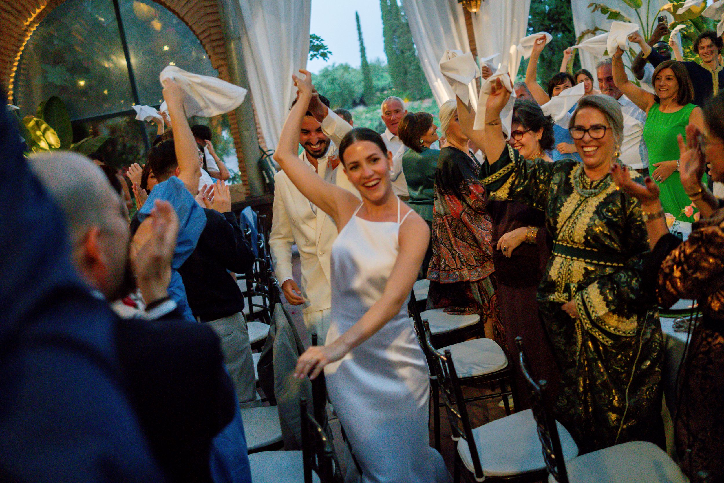 Joyful bride in white satin dress celebrates wedding ceremony exit with cheering guests raising hands.
