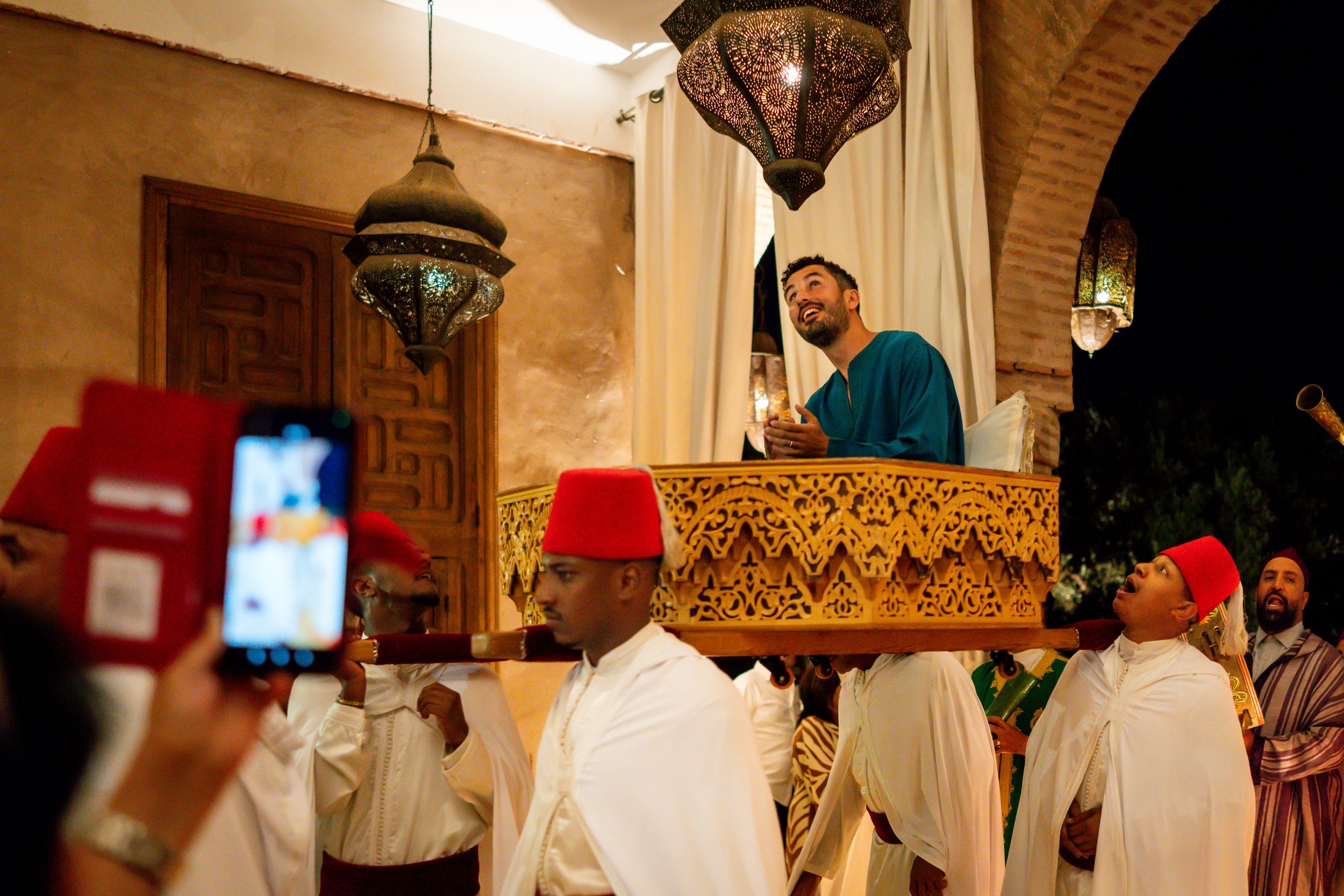 Groom carried on ornate chair during traditional Moroccan wedding celebration with men in fez hats.